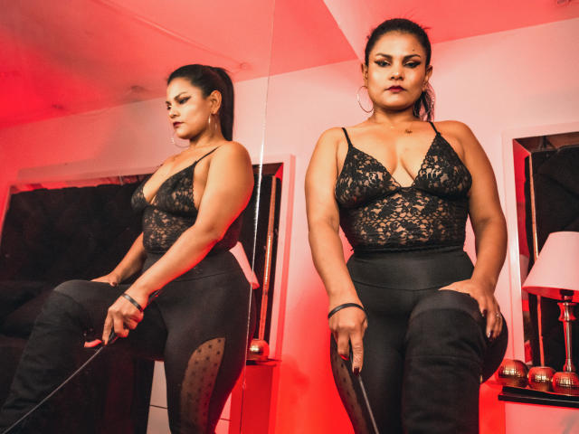 AmberColt with dark hair in a ponytail, wearing a black lace bodysuit and high-waisted black pants, standing in front of a mirror, reflecting her confident, sexy pose.