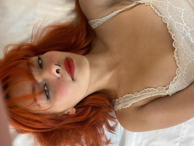 MikyValkova lying on a white bed, wearing a white lace bra, with red hair, nose ring, and red lipstick, showing her tits and a slight blush on her face.