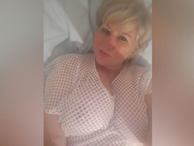 AndreaTay with short blonde hair, smiling, wearing a white mesh dress, lying on a bed, her small tits visible through the fabric, soft light on her face.
