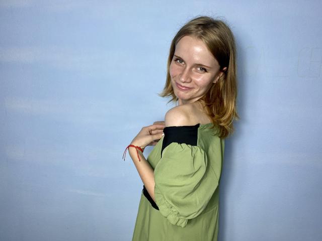 MargaritaKis with small tits, green off-shoulder dress, and red bracelet, smiling, looking over shoulder, blonde hair, light skin, blue background.