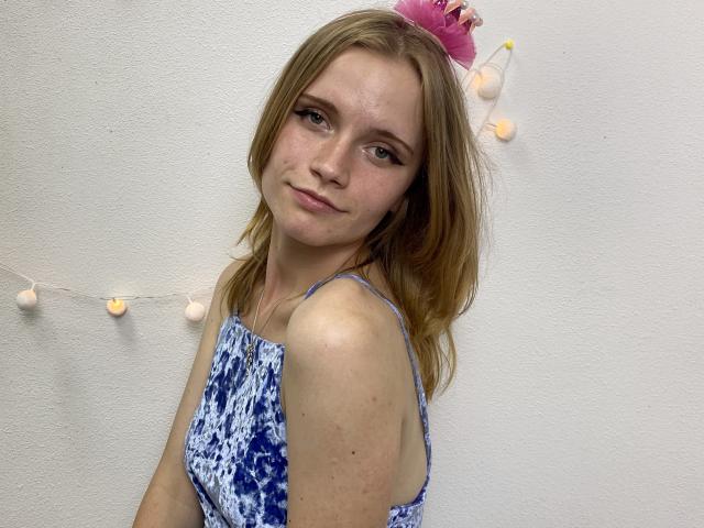 MargaritaKis with blonde hair, wearing a blue and white patterned dress, pink flower in hair, and string lights, smiling slightly, showing her small ass and cute face.
