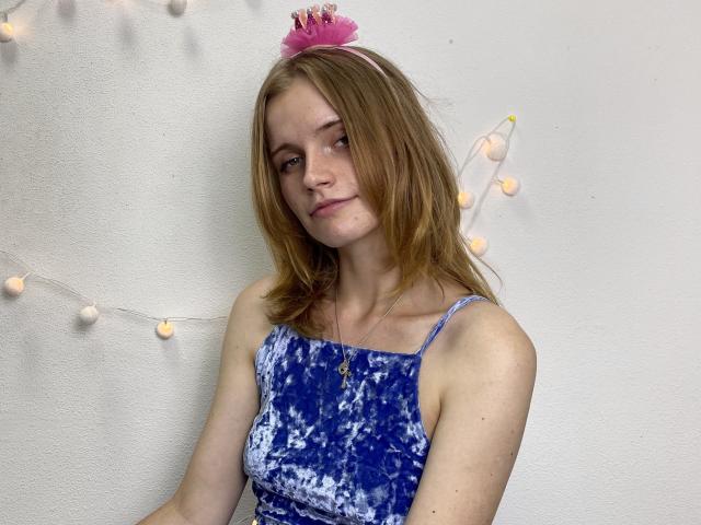 MargaritaKis with light brown hair, wearing a blue tie-dye dress, pink flower headband, and necklace, sitting against a white wall with string lights, looking slightly bored.