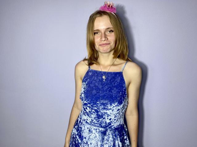 MargaritaKis avec une robe en velours bleu, une tiare rose, des cheveux bruns, de petites seins, une silhouette mince, une peau pâle, un sourire discret, un collier avec une croix, debout, fond simple, décontractée, mignonne..