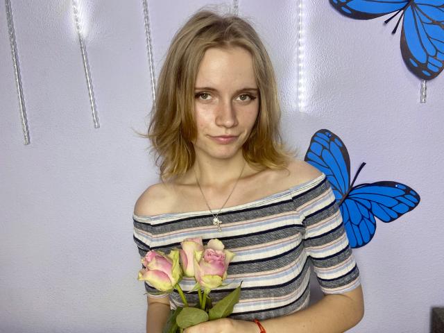 MargaritaKis, avec ses cheveux blonds, porte une chemise rayée à épaules dénudées, tient des roses roses, des autocollants de papillons bleus sur un mur blanc, un léger sourire, un collier, un bracelet rouge, une peau claire, de petites seins..