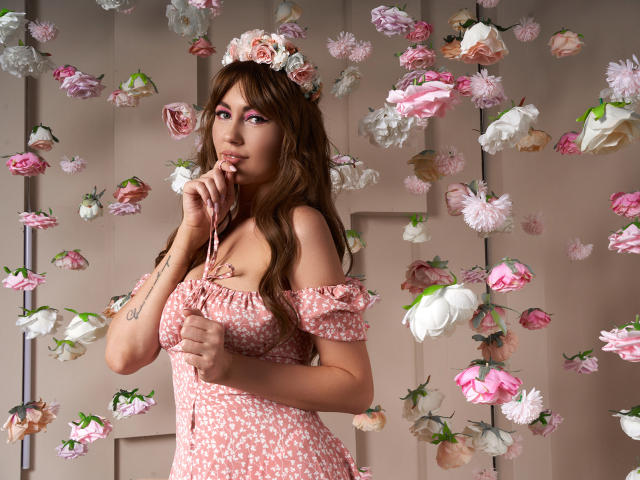 NinaTender with brown hair, pink floral dress, off-shoulder top, flower headpiece, finger to mouth, floral wall, pink and white petals, tattoo on left wrist, cute, sexy.