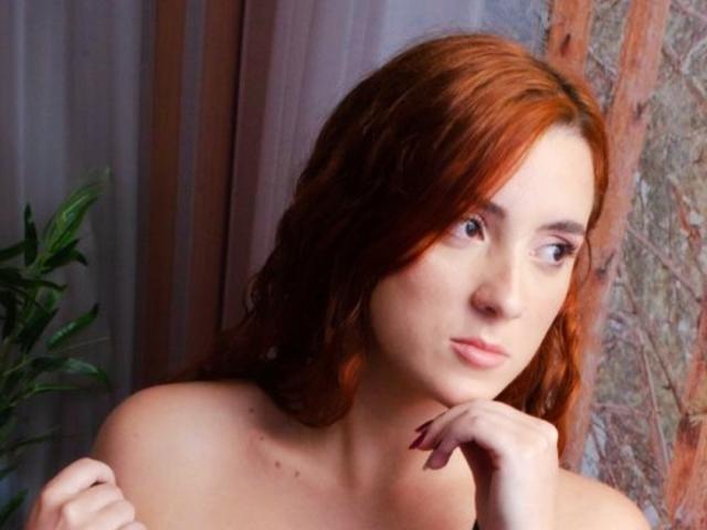 Clarice Mallor, con cabello rojo, mirando hacia un lado, tiene la barbilla apoyada en la mano, mostrando una expresión pensativa, y hombros desnudos con pequeñas marcas rojas en el pecho..