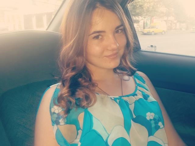 Una donna con capelli castano ricci, vestita con un abito blu a fiori, è seduta in un'auto, sorridendo leggermente, con la luce del sole che illumina il suo viso, mettendo in risalto la sua pelle morbida e la sua espressione delicata..
