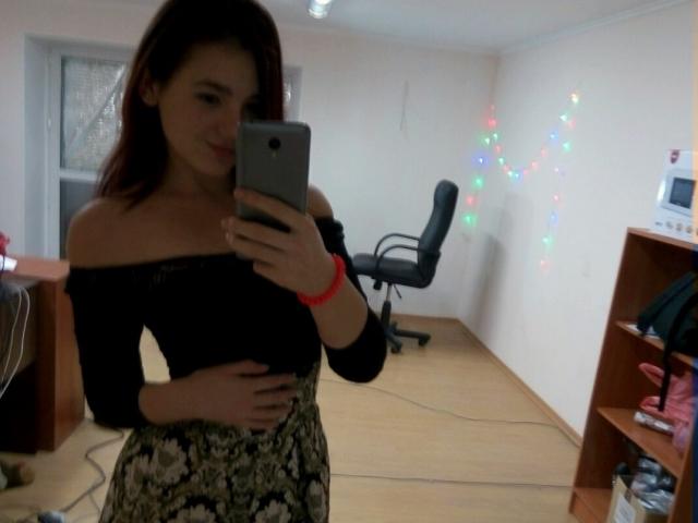 NonStop with dark red hair, black off-shoulder top, and floral skirt, holding phone, showing small tits, smiling, in a room with chair and colorful string lights.