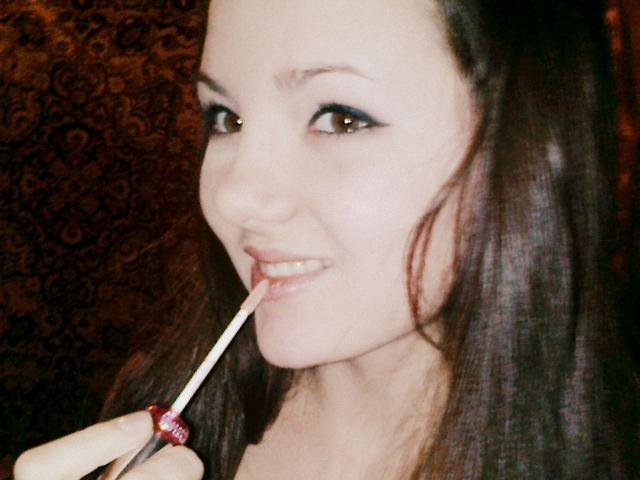 NonStop with dark brown hair, slightly smiling, using a white and red makeup brush on her pinkish lips. She has light skin and brown eyes.