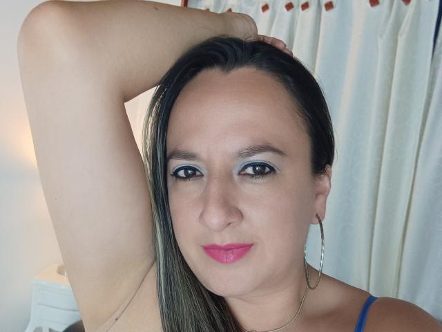CharlizeLegs ha un viso carino con rossetto rosa, occhi azzurri e capelli neri, alza un braccio, mostrando piccoli seni, indossa una maglietta blu e orecchini a cerchio..