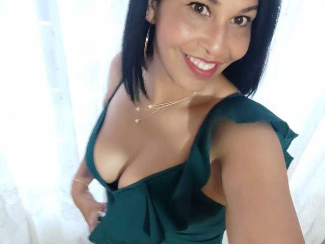 Shanon, uma mulher madura com cabelo escuro, sorrindo, vestindo um vestido verde com um decote profundo que mostra seus seios, usando um colar de ouro e brincos de argola, tirando uma selfie..