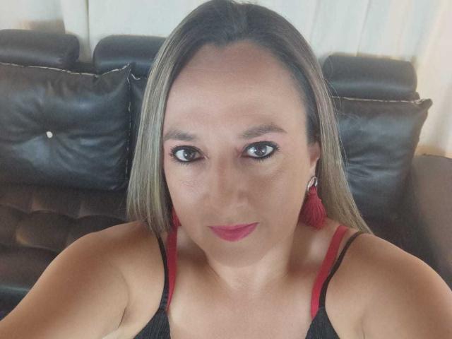 CharlizeLegs has medium brown skin, long straight brown hair, red lipstick, black tank top with red straps, and red tassel earrings, sitting on a black leather couch.