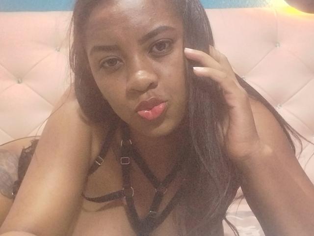 TiffanySammy with dark skin, long black hair, and pouty pink lips, wearing black strappy lingerie, touching her head with a hand, looking into the camera.