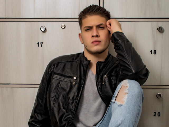 SebastiianMaxime, mit kurzen, braunen Haaren, trägt eine schwarze Lederjacke und zerrissene blaue Jeans und sitzt an einer Wand aus Holzschränken, die Hand auf dem Kopf, und wirkt leicht gelangweilt..