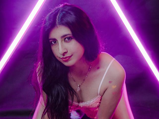 EmmaXWilliams, con lunghi capelli scuri, una maglia di pizzo rosa e una collana, guarda la fotocamera. Sullo sfondo, luci triangolari rosa neon. Trucco leggero, un sorriso carino, un leggero rossore sul seno..