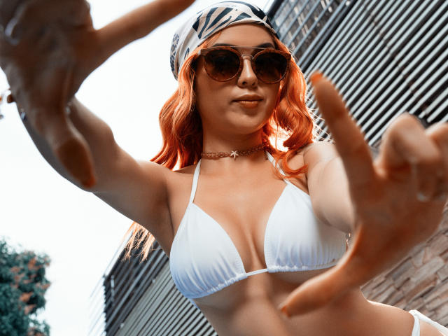 MaisyWarner with orange hair, white bikini, and sunglasses, showing off her tits in a low-angle shot, reaching forward with her hands. She's wearing a zebra-pattern cap and choker.