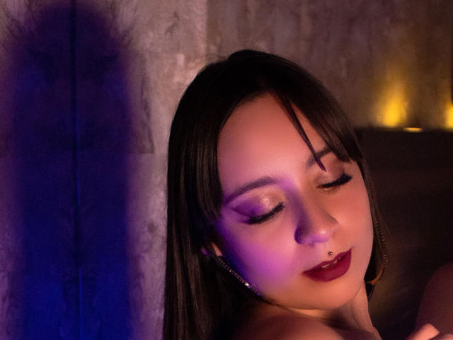 AnnPeeks, con cabello oscuro, ojos cerrados, párpados morados, lápiz labial rojo y aretes, tiene una iluminación suave en tonos morados y azules en su rostro. Está ligeramente apoyada contra una pared con textura..