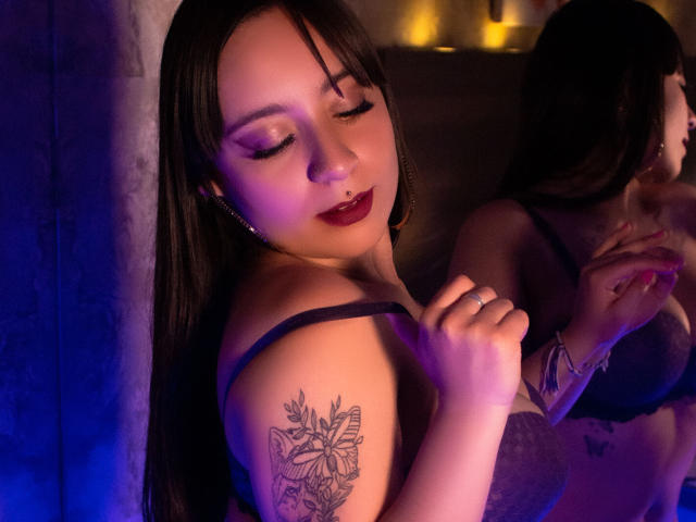 AnnPeeks, com cabelo escuro, sombra roxa e batom vermelho, segurando uma alça preta, mostrando uma tatuagem de borboleta no braço, em um quarto iluminado em tons de roxo, vestindo um sutiã preto..