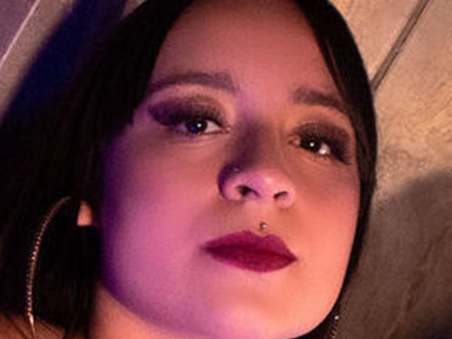 AnnPeeks tiene el cabello castaño oscuro, sombra de ojos morada, un pequeño piercing en la nariz, lápiz labial rojo, y lleva grandes aros en una imagen con poca luz y tonos morados..