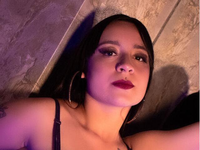 A AnnPeeks tem cabelo escuro, brincos grandes, um piercing no nariz e batom roxo. Ela está olhando para cima com um leve sorriso, vestindo uma blusa preta de alça fina. Seus seios estão visíveis..