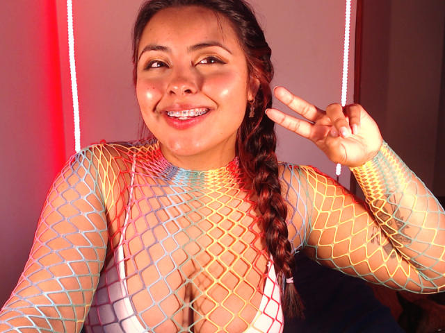 AngelaRoggers with shiny skin, smiling, wearing a rainbow fishnet top, exposing her large tits, and a white bra, making a peace sign, with her brown braid.
