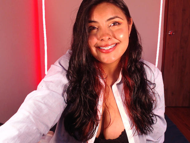 Angela Roggers, avec de longs cheveux noirs ondulés, souriante, portant une chemise blanche déboutonnée laissant apparaître un soutien-gorge en dentelle noire, et un éclairage rouge en arrière-plan. Ses seins sont visibles..