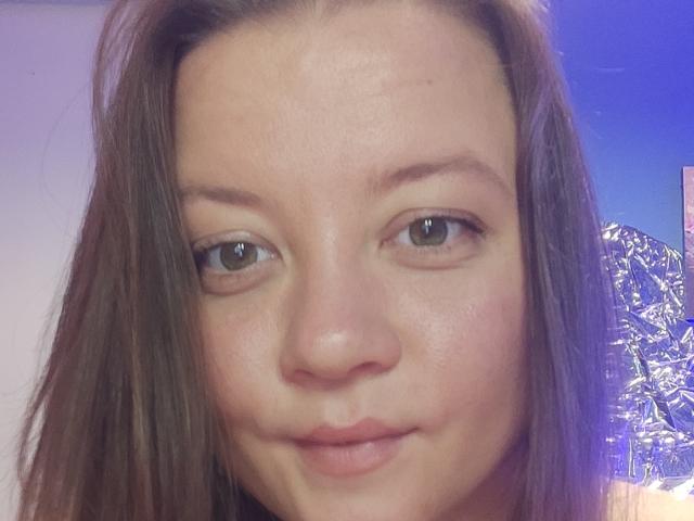 Het gezicht van NessLulu, met bruine ogen, steil bruin haar, lichtrode huid en een subtiele grijns. Haar kleine neus en roze lippen zijn zichtbaar. Blauw licht op de achtergrond..