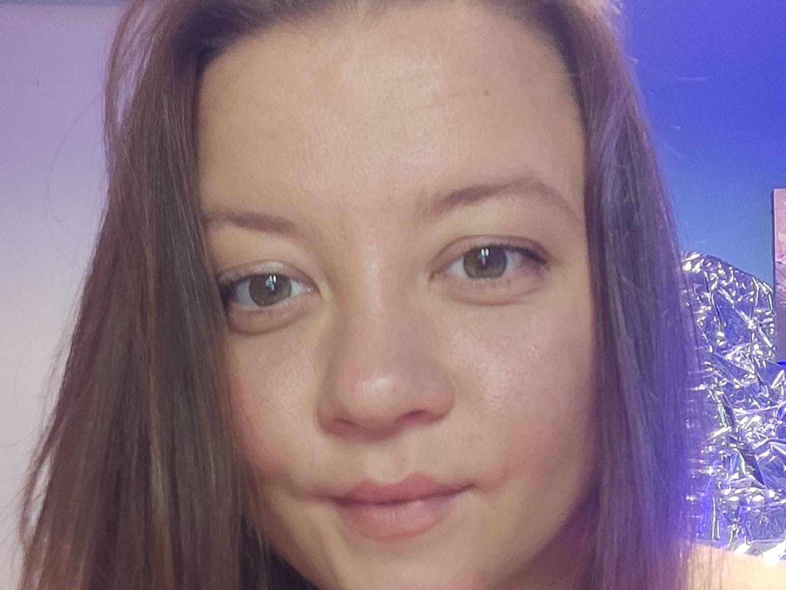 O rosto de NessLulu, mostrando olhos castanhos, cabelo castanho liso, pele levemente avermelhada e um sorriso discreto. Seu pequeno nariz e lábios rosados são visíveis. Luz azul ao fundo..