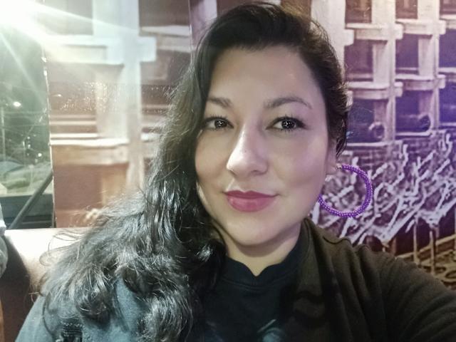 Sakoto Dika, avec ses longs cheveux noirs, ses boucles d'oreilles violettes et son rouge à lèvres rose, sourit à l'appareil photo, portant une chemise noire. Elle a un visage mignon et un petit nez..