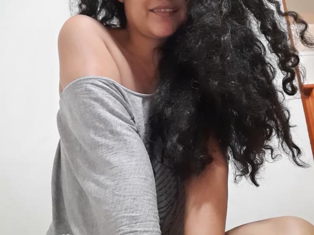 Xuuley, con lunghi capelli ricci neri, sorride, indossa una maglia grigia a spalle scoperte, mostrando la spalla nuda e la parte superiore del petto, ed è seduta con le gambe piegate..