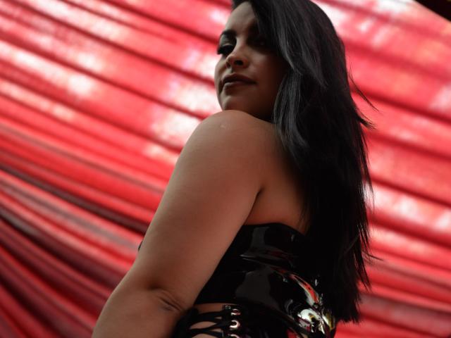 PaulinaXHornyX with long black hair, looking back, smiling, wearing a shiny black strapless top, exposing her shoulder and a small tattoo, with red textured background.