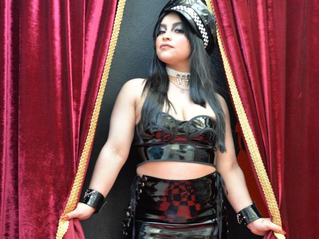 PaulinaXHornyX with black latex outfit, strapless top showing tits, choker, and skirt with checkered red pattern, holding red velvet curtains, black wall in background.