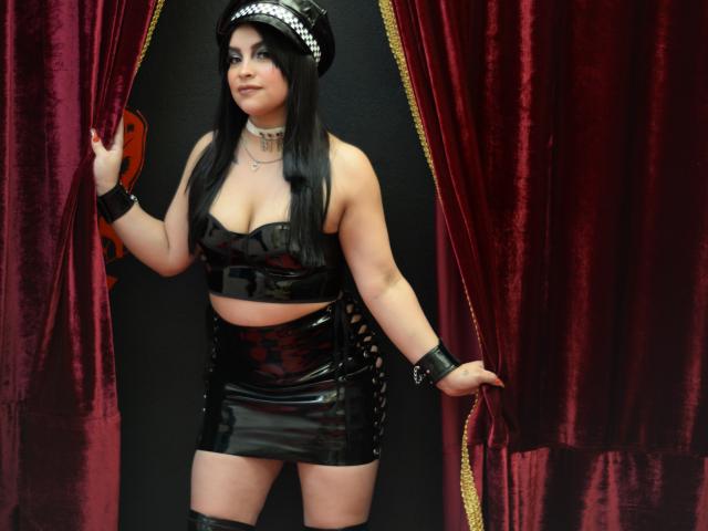 PaulinaXHornyX, vêtue d'une combinaison en latex noir avec des découpes, a de grandes seins, porte un crop top moulant et une mini-jupe, des bottes noires montantes et une casquette de policier, et tient des rideaux rouges..