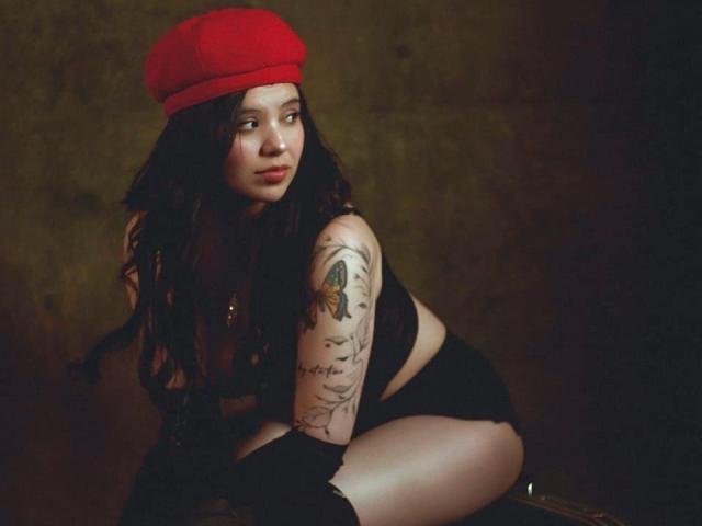 ElyMoon with long dark hair, red beret, black top, and black shorts, tattooed arm, crouching, looking sideways, dark background, sexy, pale skin, soft lighting, slight pout.