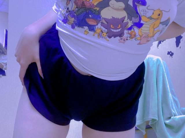 Rubbi's ass in dark blue shorts, hand pulling up the waistband. Wearing a white Pokémon shirt, visible from waist down. Blue towel hanging in the background.
