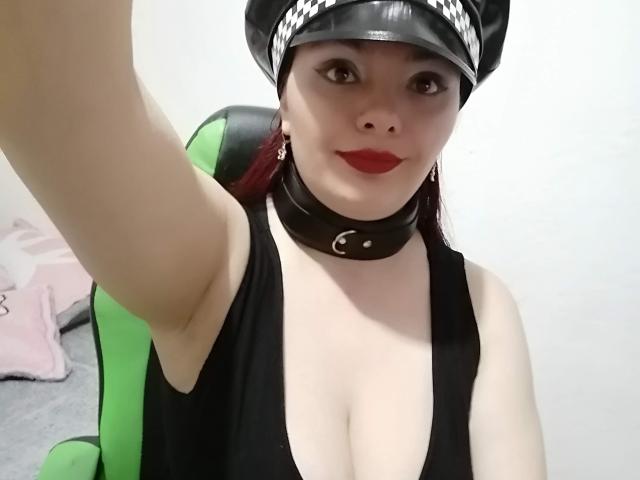 Katherin, con rossetto rosso, berretto di pelle nera, collana e profonda scollatura in una canotta nera, alza un braccio per un selfie, mostrando i suoi seni e l'ascella..