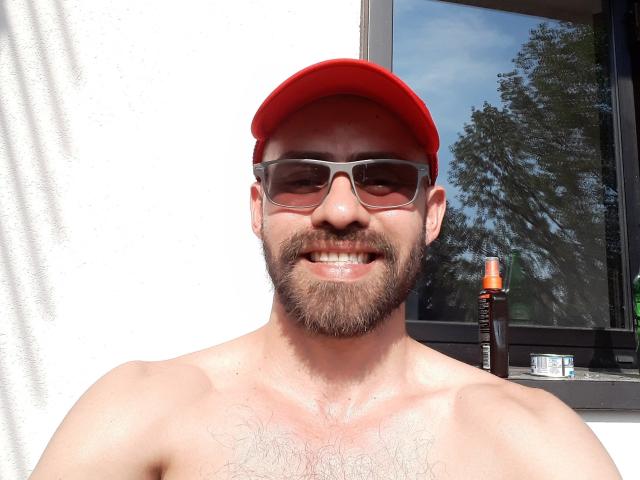 DavidHott69 zonder shirt, met een rode pet, zonnebril, baard en snor, lachend. Hij heeft een behaarde borst, bruine huid, en een fles zonnebrandcrème op een vensterbank aan de rechterkant..