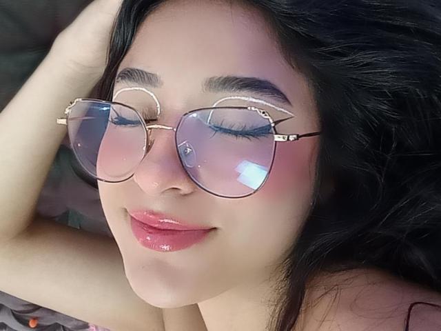 ValeriaClark has big-ass glasses with reflective lenses, dark eyebrows, blue eyeshadow, and a shiny red lipstick smile. Her black hair is slightly messy.