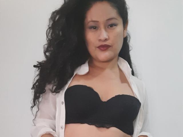EmilyJensenn with long black hair, white crop top, black sequin shorts, white thigh-high socks, sitting on bed, showing her ass, small tits, and a cute face, in a bedroom.