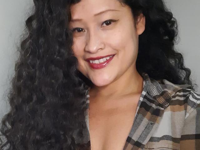 Xuuley tem cabelos pretos encaracolados, batom vermelho e um leve sorriso, mostrando seios à mostra com um tom de pele bronzeado, olhando para cima com olhos escuros. A marca d'água "u/hellhoney" é visível..