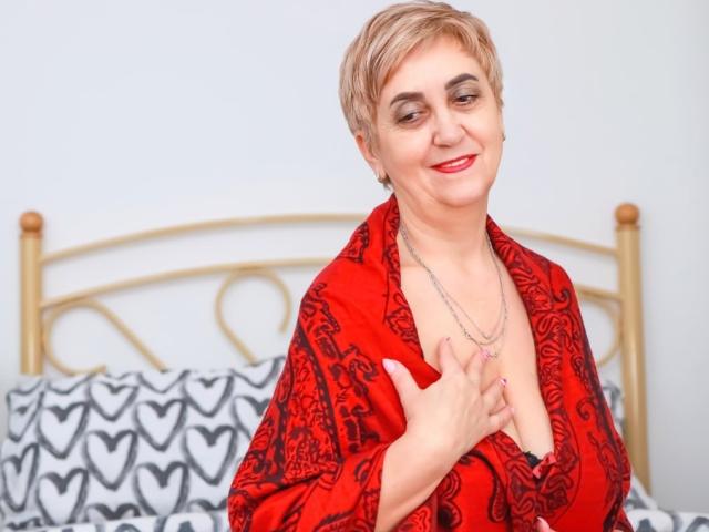 MadamMargo with short blonde hair, red patterned robe, smiling, red lipstick, holding robe open, showing cleavage, silver necklace, standing in front of a bed with heart-patterned sheets.