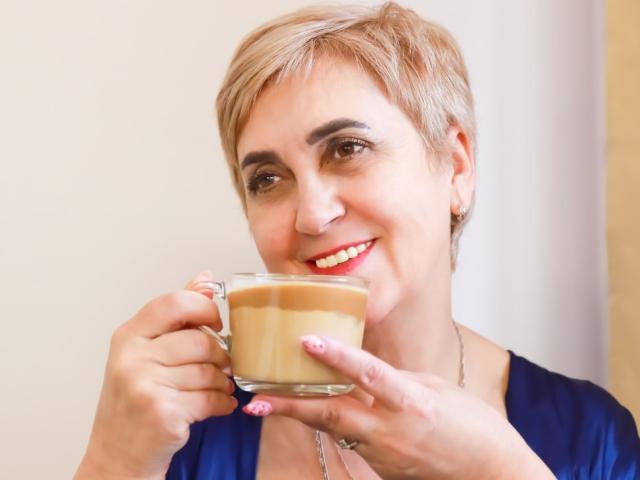 MadamMargo with short blonde hair, blue shirt, red lipstick, holding a cup of coffee, smiling, pink nail polish, and a silver necklace, looking happy.