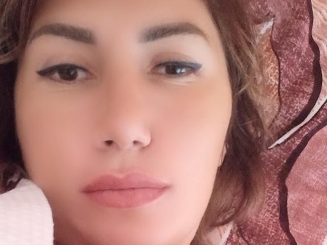 RoxolanaSexy's Gesicht mit brauen Augenbrauen, blauem Eyeliner und pinkem Lippenstift. Sie hat ein Nasenpiercing und braune, wellige Haare und blickt leicht zur Seite, mit einem neutralen Ausdruck..
