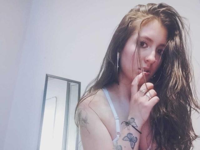 LexySmithX met lang bruin haar, die haar vinger bijt, gekleed in witte lingerie, met tatoeages zichtbaar op haar linkerarm en hand, draagt witte oordopjes, in een zacht verlichte kamer..