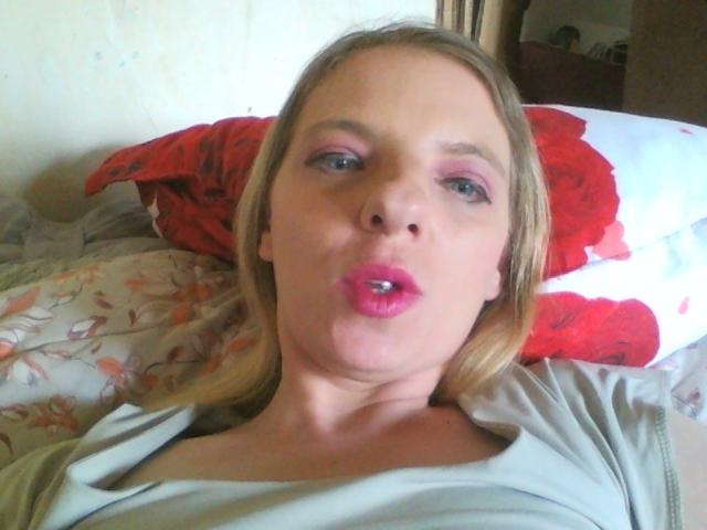 BlondyMarianne's gezicht, met roze lippen die licht open zijn, roze oogschaduw, blond haar, een grijze trui, een rood-witte kussen, een bloemenprint bedlaken, licht opgezwollen wangen, een casual selfie in de slaapkamer..