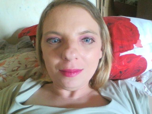 BlondyMarianne, avec ses yeux bleus, son rouge à lèvres rose et son fard à paupières rose, est allongée sur un lit avec un oreiller rouge, vêtue d'une chemise blanche, et elle a un léger sourire tout en regardant l'appareil photo..