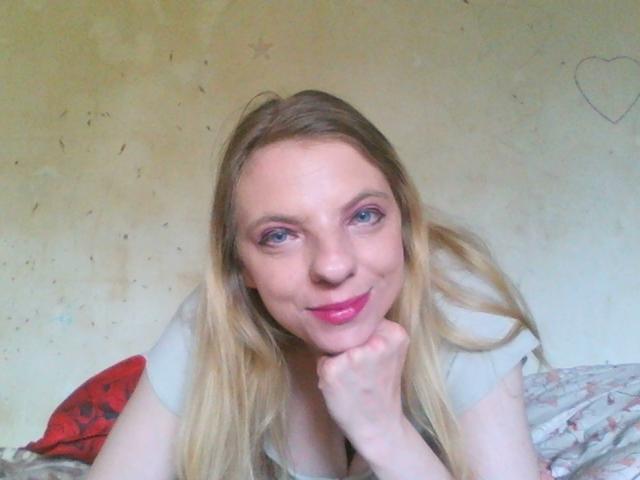 BlondyMarianne mit rosafarbenem Lippenstift, blauen Augen und langen blonden Haaren, lächelnd, mit dem Kinn in der Hand, trägt ein graues Oberteil und zeigt ihre Brüste leicht..
