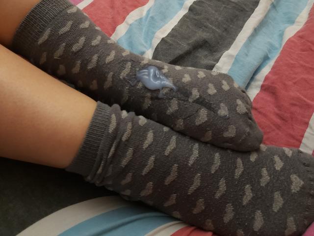 Aline, con las piernas cruzadas, lleva calcetines grises con un estampado, y en el calcetín derecho se ve una pulsera de plástico azul. Los calcetines tienen pequeños diseños grises que se repiten..