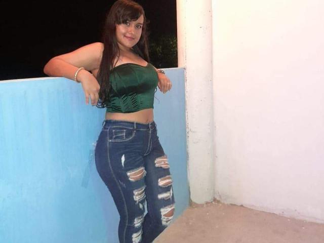 Elsa Santos mit langen, braunen Haaren, einem grünen Sam-Oberteil und zerrissenen, blauen Jeans, lehnt an einer blauen Wand, lächelt und zeigt ihre kleinen Brüste und ihre schlanke Taille..