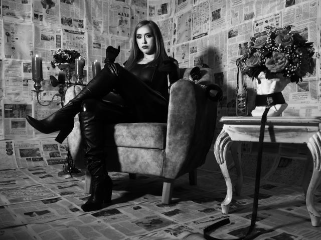 VenusBarret with dark lipstick, sitting in a chair, wearing a black top and thigh-high leather boots, legs crossed, arms resting on chair arms, black roses on table.